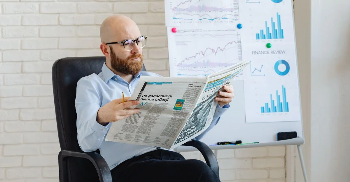 Homme chauve avec une barbe lisant un journal dans un bureau avec des graphiques financiers sur un tableau blanc.