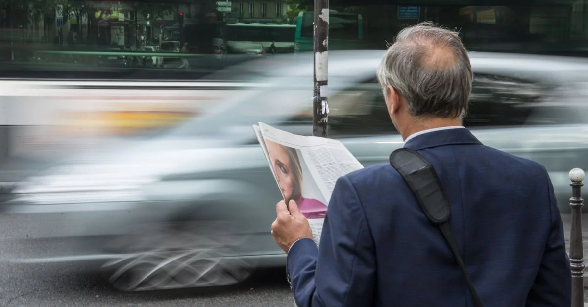 Un homme âgé lit un journal dans une rue animée de la ville avec un flou de mouvement dans la circulation.