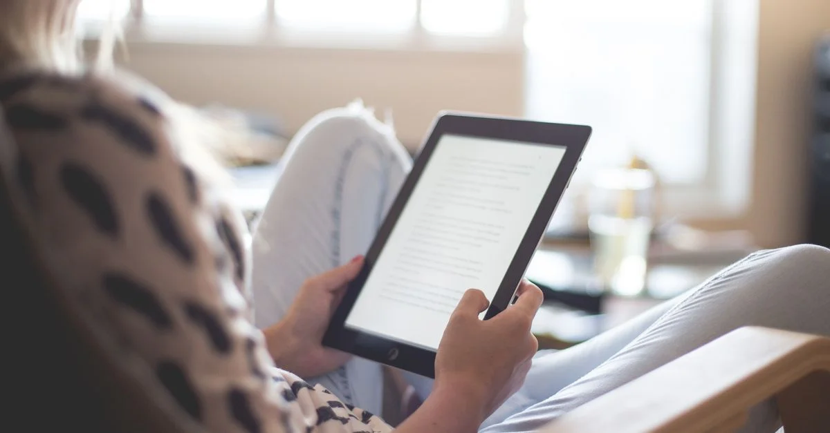Une femme apprécie la lecture sur une tablette, assise confortablement dans un salon moderne.