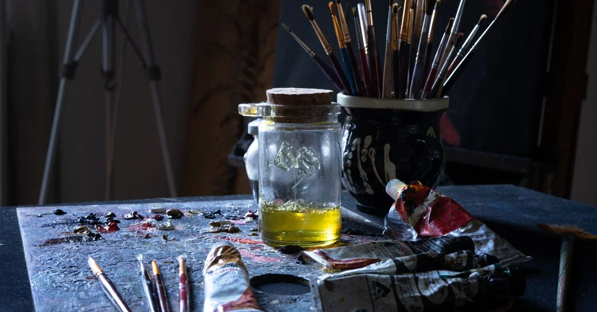 Une table d'artiste en désordre remplie de tubes de peinture, de pinceaux et d'un pot en verre.