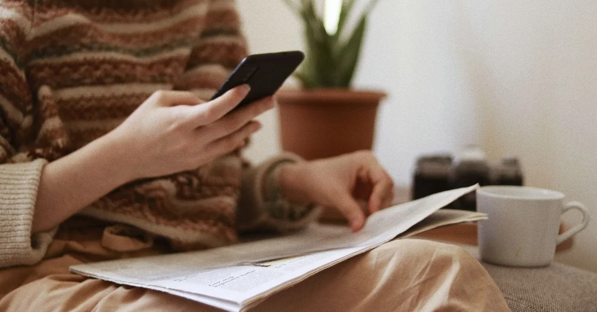 Une femme profite de son temps libre à consulter son smartphone tout en lisant un journal à l'intérieur.