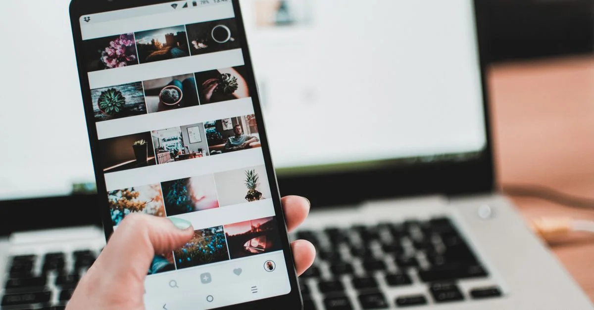 Une personne utilise un smartphone pour consulter les réseaux sociaux devant un ordinateur portable, soulignant la technologie moderne et le style de vie.